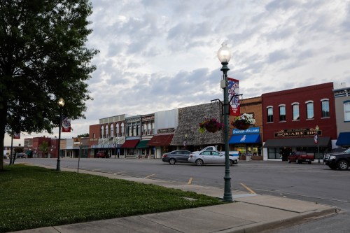 A quiet downtown street with shops, lampposts, and a cloudy sky in the background. Green grass in the foreground.