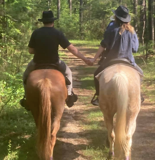 Two people on horseback hold hands while riding down a forest path, surrounded by greenery.