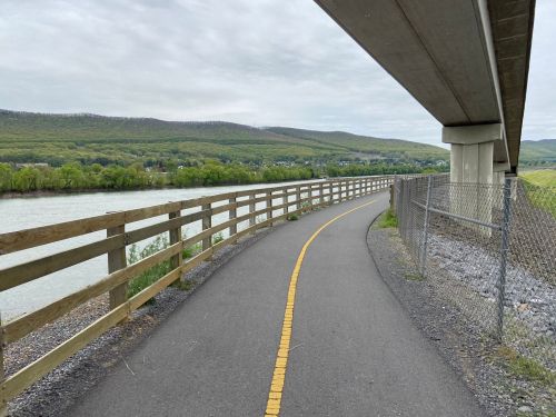 A paved path alongside a river, bordered by a wooden fence and a chain-link fence, with hills in the background.