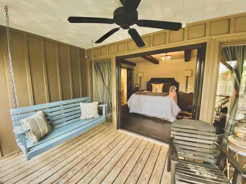 A cozy porch with a blue swing and wooden furniture, leading to a stylish bedroom with a large bed and warm decor.