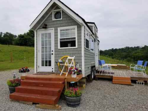 A small, charming tiny house on wheels with a wooden deck, surrounded by greenery and colorful flower pots.