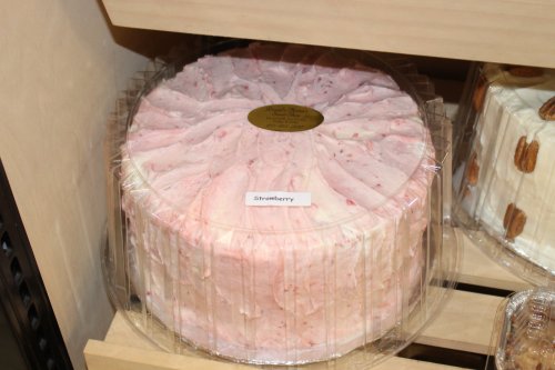 A round strawberry cake with pink frosting, displayed in a clear plastic container on a shelf.