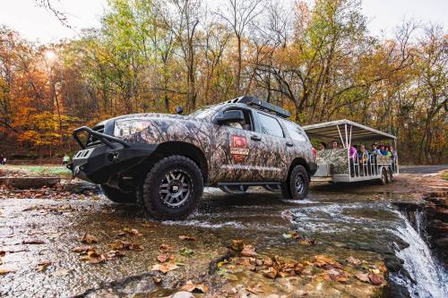 A camouflaged SUV parked by a stream, with a trailer carrying people in a forested area during autumn.