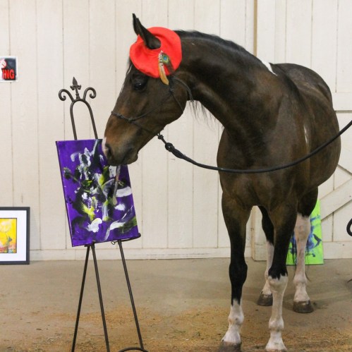 A horse wearing a red hat stands beside a colorful painting in an indoor setting.