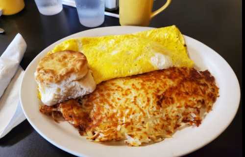 A plate of breakfast featuring a yellow omelet, crispy hash browns, and a biscuit, served on a black table.