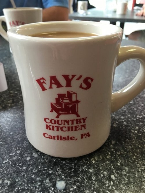 A white coffee mug with "Fay's Country Kitchen, Carlisle, PA" printed in red, filled with coffee on a table.