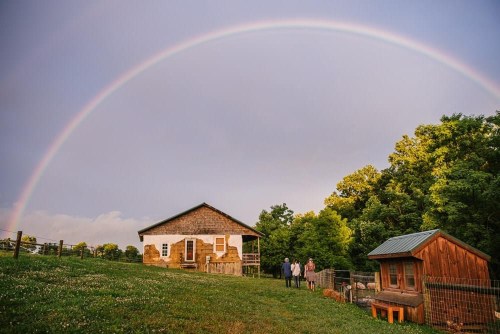 A rainbow arcs over a rustic house and a small animal shelter, with three people standing on the grassy lawn.