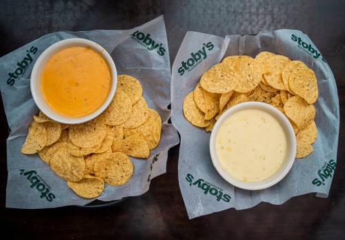 Two bowls of cheese dip, one orange and one white, surrounded by tortilla chips on branded paper.
