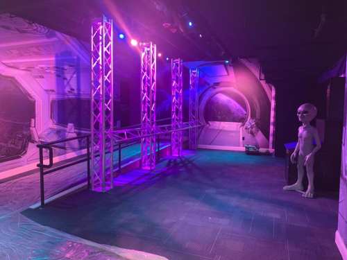 A dimly lit space with purple lights, featuring a futuristic backdrop and a life-sized alien figure standing beside a railing.