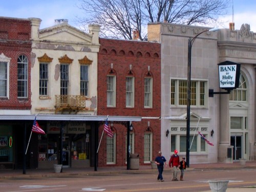 Historic buildings line a street in Holly Springs, with American flags and pedestrians walking by.
