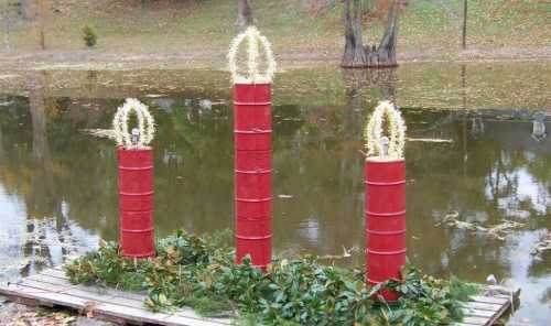 Three large red candles with decorative crowns, surrounded by greenery, set by a calm pond in a park.