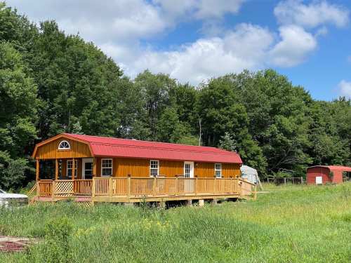 A bright yellow cabin with a red roof sits on a grassy field, surrounded by trees and a blue sky with clouds.