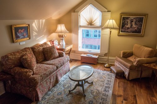 Cozy attic living space with a floral sofa, armchair, coffee table, and large window letting in natural light.