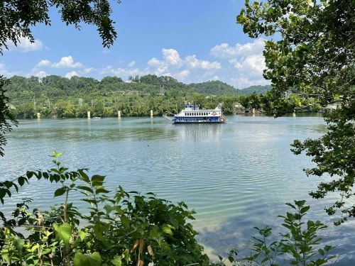 A ferry boat glides across a calm river, surrounded by lush greenery and blue skies with scattered clouds.
