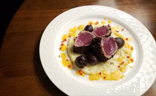 A plate of seared tuna on creamy grits, garnished with roasted grapes and a drizzle of sauce.