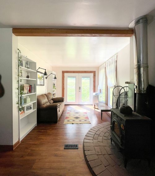 Cozy living space with a brown couch, bookshelves, and a wood stove, featuring large windows and a door to the outdoors.