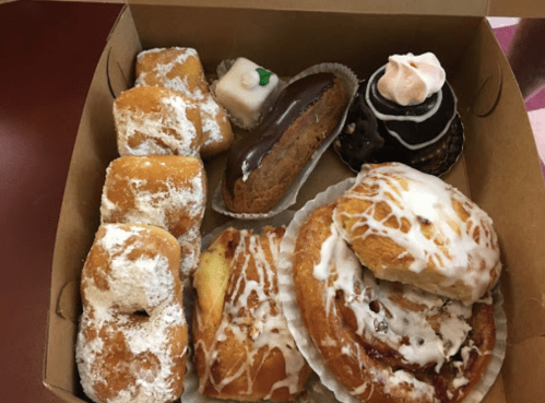 A box filled with various pastries, including cinnamon rolls, donuts, and small cakes, dusted with powdered sugar.