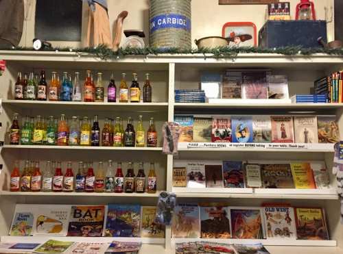 A shelf filled with colorful soda bottles and various books, including children's stories and local history.