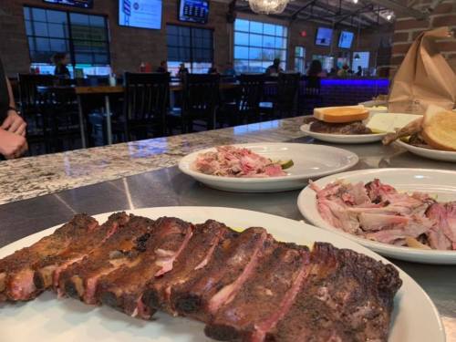 A plate of sliced barbecue ribs in a restaurant, with additional plates of pulled pork and sides in the background.