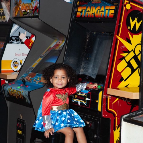 A young girl dressed as Wonder Woman plays an arcade game in a colorful gaming area.