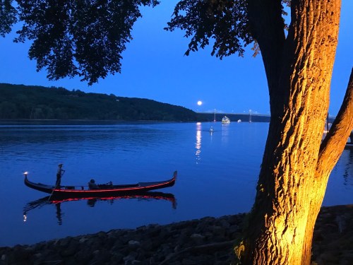 A serene river scene at dusk, featuring a gondola on the water and a tree illuminated by warm light.