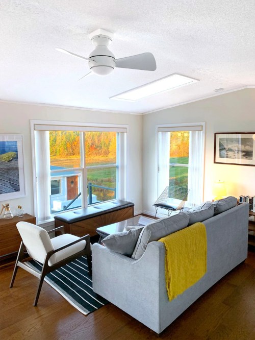 Cozy living room with a gray sofa, yellow throw, and large windows showcasing a colorful autumn landscape outside.