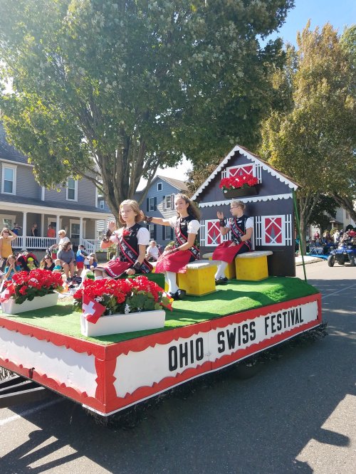 Three girls in traditional Swiss dresses sit on a decorated float at the Ohio Swiss Festival, surrounded by flowers.