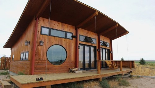 A modern wooden house with large windows, a circular window, and a spacious deck in a rural setting.