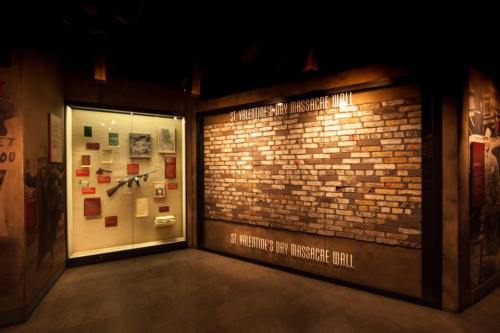 A dimly lit exhibit featuring the St. Valentine's Day Massacre Wall and a display case with historical artifacts.