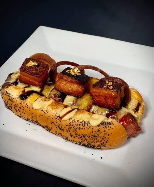 A gourmet hot dog topped with crispy pork belly, drizzled with sauce, and garnished with gold flakes on a white plate.