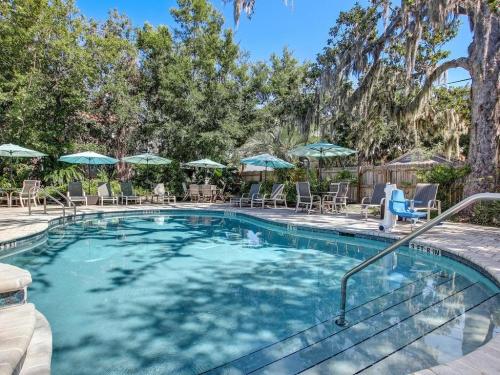 A serene pool area surrounded by trees, lounge chairs, and umbrellas under a clear blue sky.
