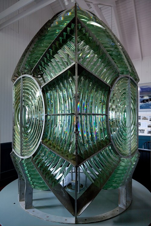 A large, intricate glass lighthouse lens with multiple curved panels, reflecting light in various colors.