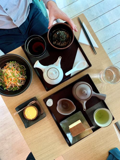 A top-down view of a dining table with two trays featuring tea, noodles, a dessert, and a small dish of seaweed.