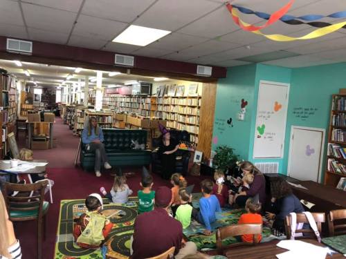 A children's storytime in a library, with kids sitting on a rug and adults reading on a couch surrounded by bookshelves.