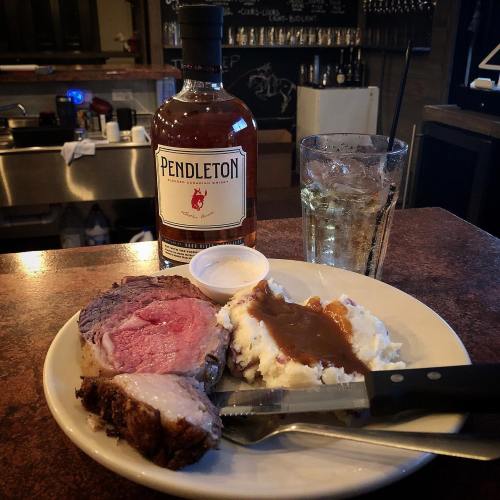 A plate with prime rib, mashed potatoes, and gravy, alongside a bottle of Pendleton whiskey and a glass of mixed drink.