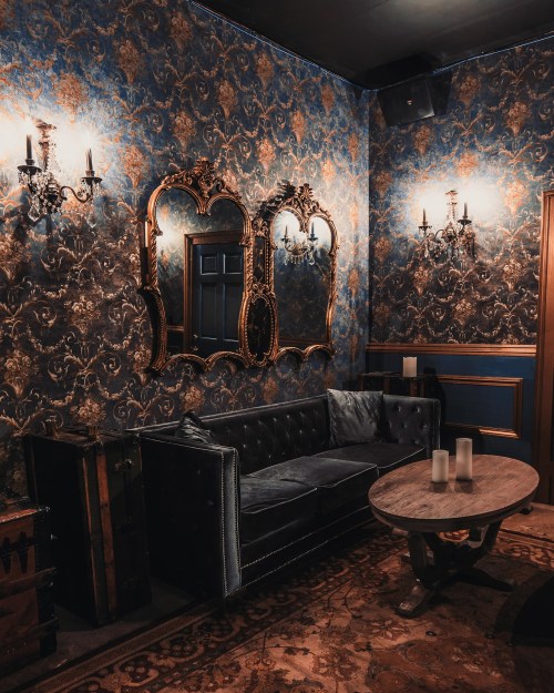 A cozy, elegantly decorated room featuring a dark sofa, ornate mirrors, and vintage wallpaper with soft lighting.