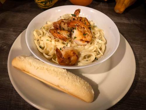 A bowl of fettuccine pasta topped with shrimp, served with a side of breadstick on a plate.