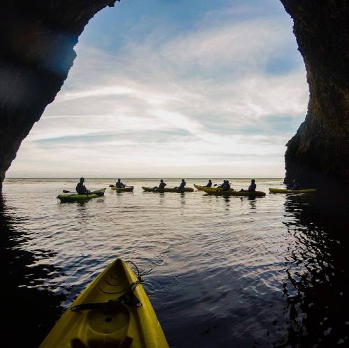 A group of kayakers paddles through a cave, with calm waters and a cloudy sky in the background.