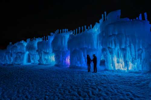 Two silhouetted figures stand in front of illuminated ice formations, glowing blue against a dark night sky.