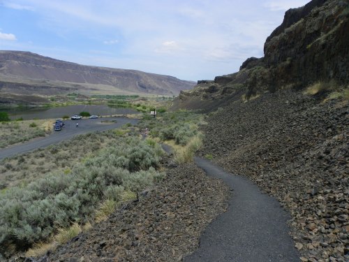 A winding path through rocky terrain leads to a parking area and lush greenery in the distance under a cloudy sky.