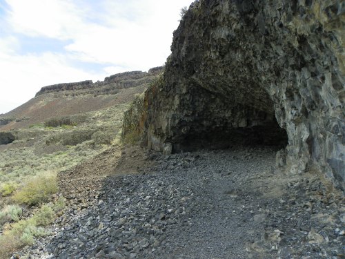 A rocky cave entrance surrounded by rugged terrain and sparse vegetation under a cloudy sky.