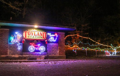 A brightly lit sign reading "Toyland Station" with colorful holiday lights in a dark, wooded area.