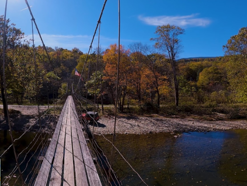 A wooden suspension bridge spans a river, surrounded by trees with autumn foliage under a clear blue sky.