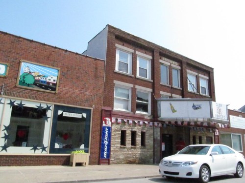 A brick building with multiple windows, featuring storefronts and a white car parked in front under a clear blue sky.