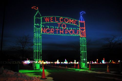 Colorful holiday lights form an archway that reads "Welcome to North Pole" against a twilight sky.