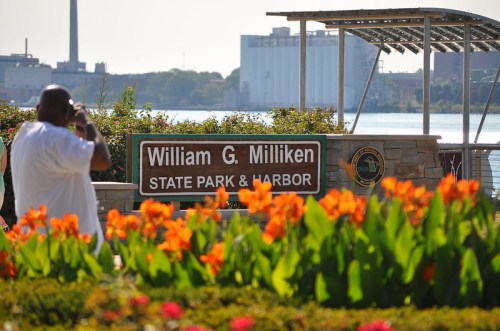 A man takes a photo near the William G. Milliken State Park & Harbor sign, surrounded by vibrant flowers.