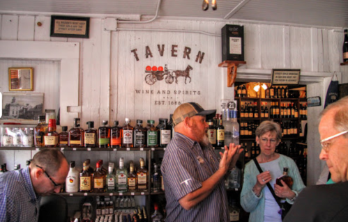 A cozy tavern interior with shelves of spirits, a bartender speaking, and patrons engaged in conversation.