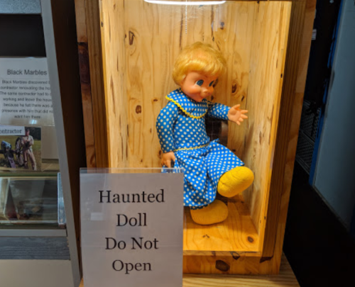 A doll in a glass display case, wearing a blue polka dot dress, with a sign reading "Haunted Doll Do Not Open."