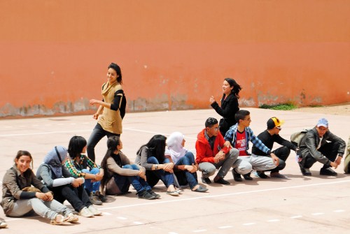 A group of young people sitting on a court, with two individuals standing and interacting playfully.
