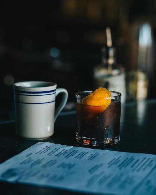 A glass of dark drink with orange peel sits next to a white mug and a menu on a table.